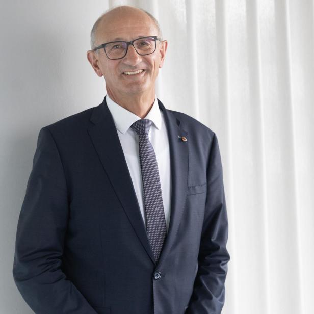 Ein älterer Mann mit Brille und Anzug steht lächelnd vor einem weißen Vorhang.