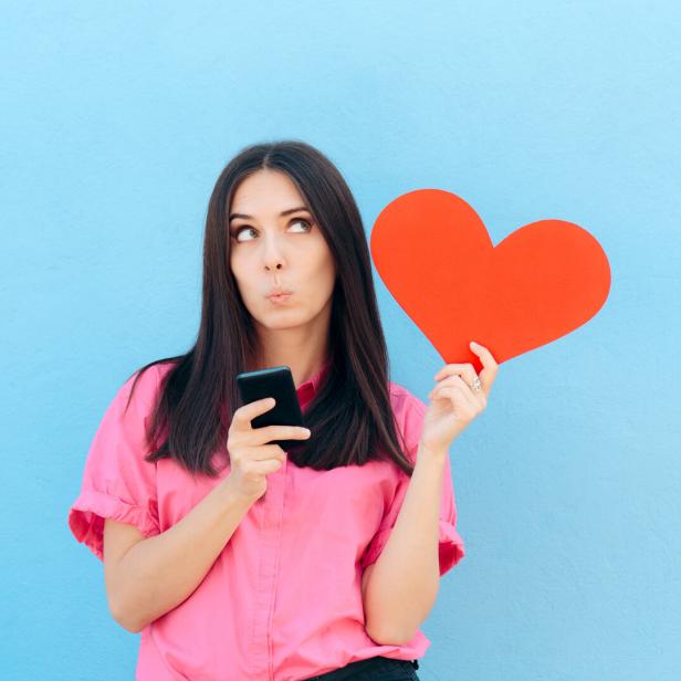 Frau in rosa Hemd hält ein rotes Herz und ein Smartphone vor blauem Hintergrund und schaut nachdenklich nach oben.