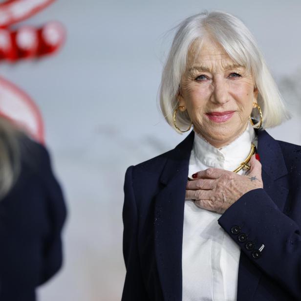 Helen Mirren lächelt in einem dunklen Blazer mit goldenen Ohrringen und einer Halskette.