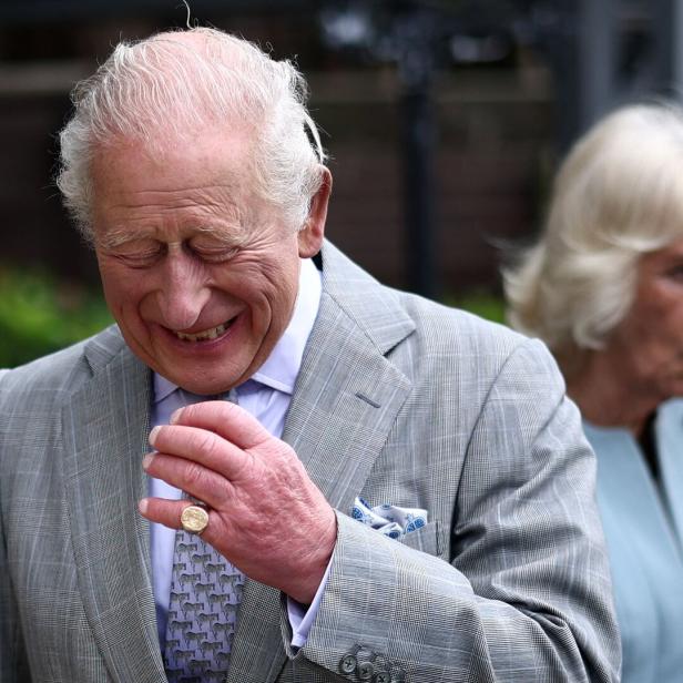 König Charles III. lächelt in einem grauen Anzug, während Camilla im Hintergrund zu sehen ist.