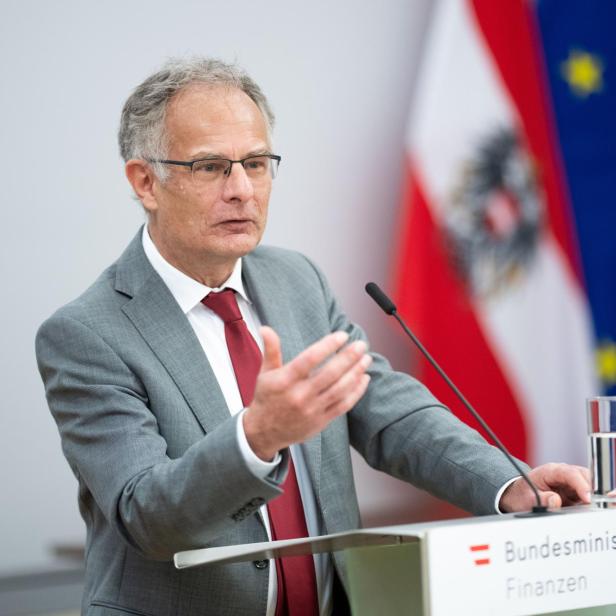 Ein Mann im grauen Anzug spricht an einem Rednerpult mit Wasserglas, im Hintergrund österreichische und EU-Flagge.