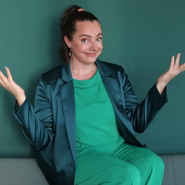 Eine Frau in grüner Kleidung und einem bläulichen Blazer sitzt mit fragenden, offenen Händen vor einem grünen Hintergrund.
