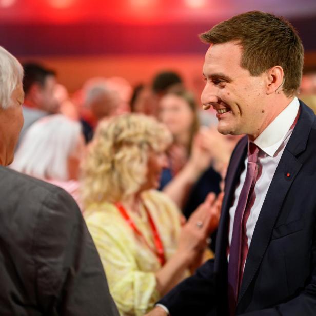 Ein Mann im Anzug lächelt und schüttelt einer anderen Person die Hand, umgeben von applaudierenden Menschen.