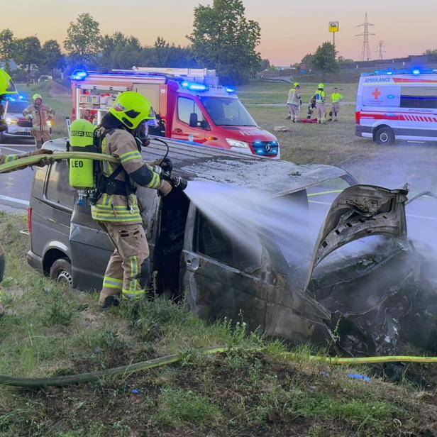 Zwei Feuerwehrleute löschen ein demoliertes Unfallfahrzeug am Straßenrand.