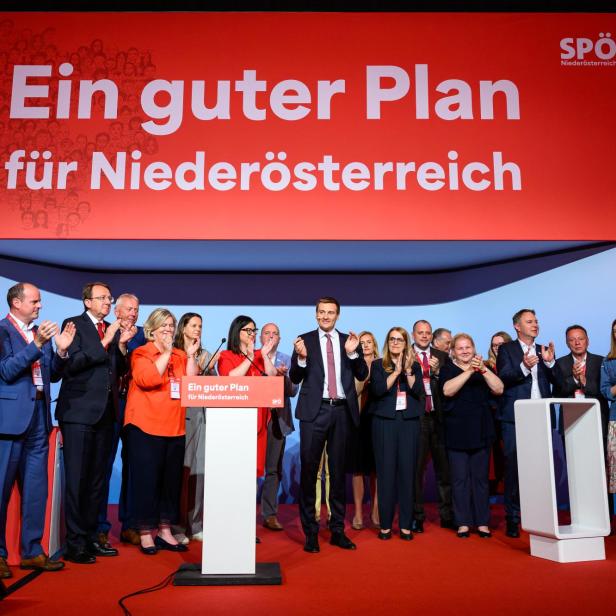 Eine Gruppe von Menschen steht auf einer Bühne vor einem großen roten Schild mit der Aufschrift „Ein guter Plan für Niederösterreich“ und applaudiert.