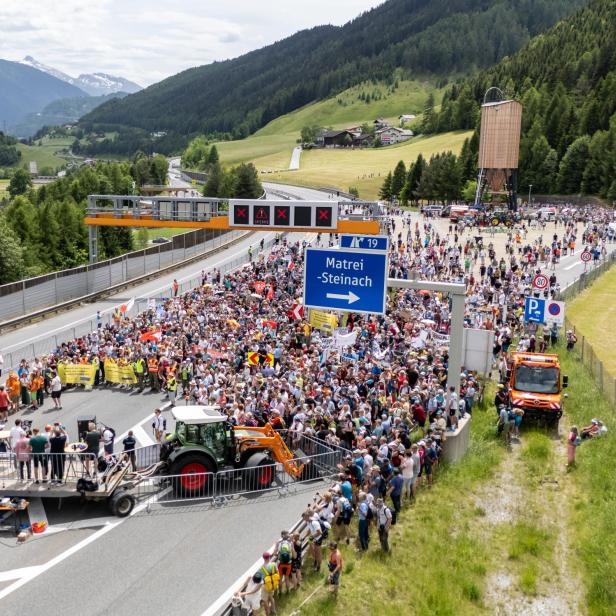 Viele Menschen versammeln sich auf einer gesperrten Autobahn in den Bergen, einige halten Transparente und Schilder.