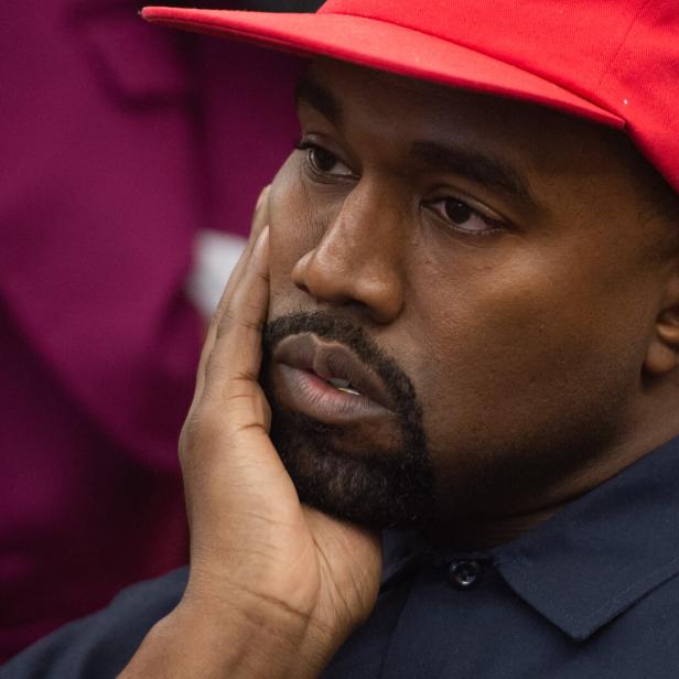 Kanye West trägt eine rote Baseballkappe und stützt nachdenklich sein Gesicht mit der Hand ab.