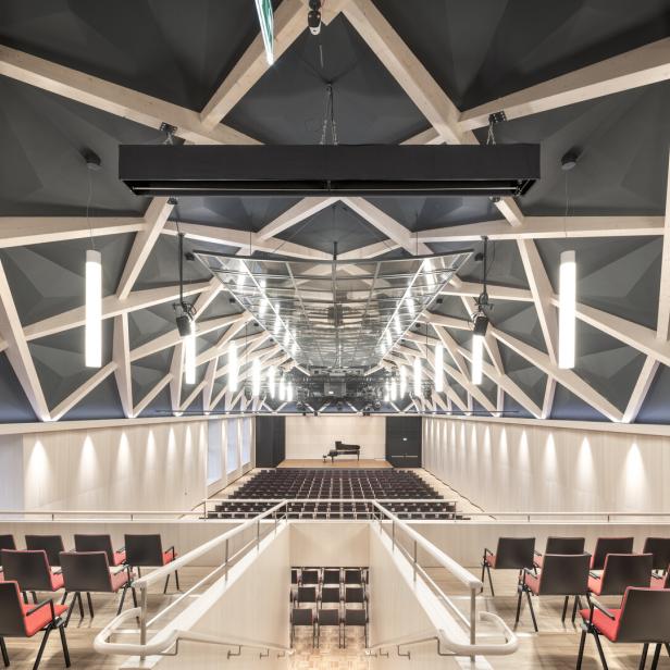 Großer moderner Konzertsaal mit geometrischer Decke, hängenden Leuchten, roten Stühlen und einem Flügel auf der Bühne.