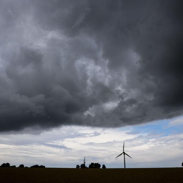 Dunkle Gewitterwolken ziehen über eine Landschaft mit Windrädern und vereinzelten Bäumen am Horizont.