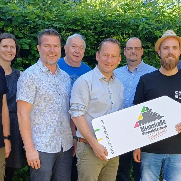 Acht Personen stehen lächelnd nebeneinander im Freien, zwei halten ein Schild mit der Aufschrift „Eisenstraße Niederösterreich“.