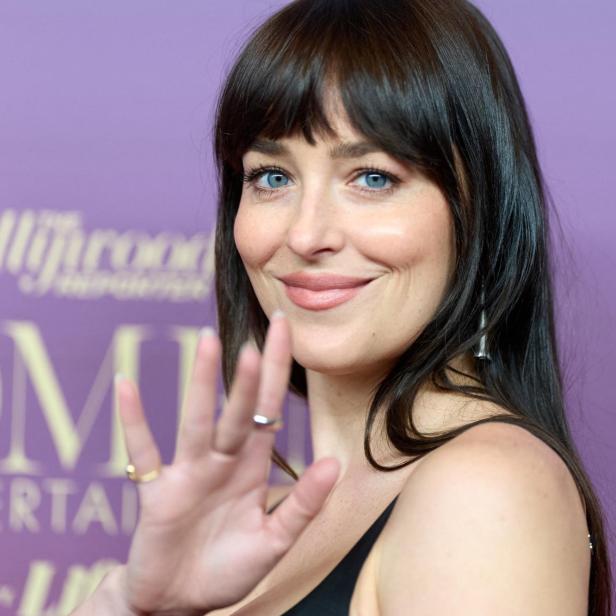 Dakota Johnson lächelt und winkt vor einem lila Hintergrund.