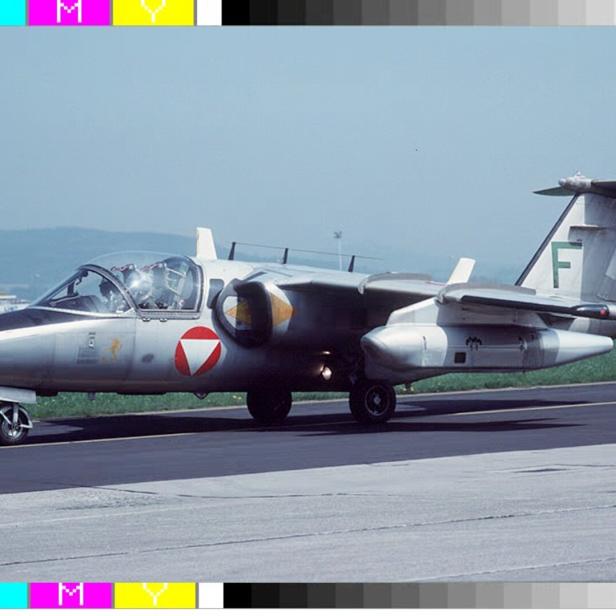 Ein silbernes zweistrahliges Militärflugzeug mit rotem Emblem steht auf einer Startbahn im Freien.