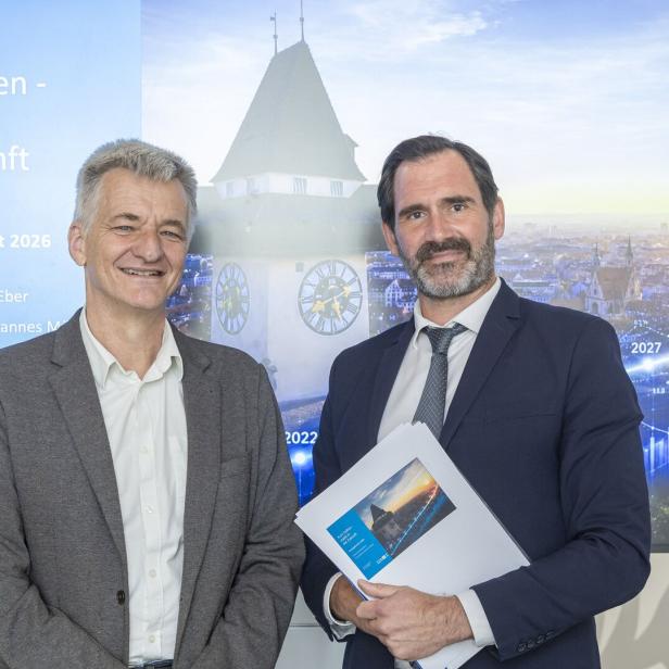 Zwei Männer im Anzug stehen vor einer Präsentation mit dem Titel „Kurs halten – stabil in die Zukunft“ und lächeln in die Kamera.