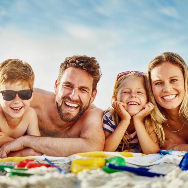 Vierköpfige Familie liegt lachend am Strand mit Strandspielzeug und Ball.