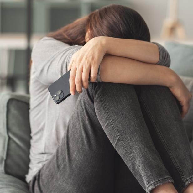 Eine Frau sitzt mit gesenktem Kopf auf einem Sofa und hält ein Smartphone in der Hand.