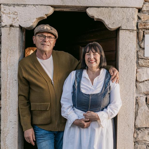 Ein kroatisches Gastgeberpaar steht lächelnd in traditioneller Kleidung vor einem Steinhaus mit dem „Local Host“-Gütesiegel an der Wand.