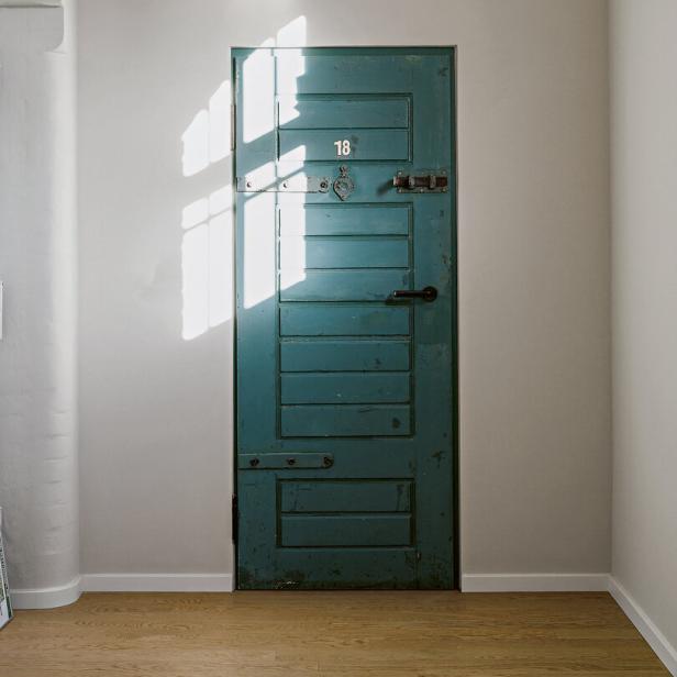 Eine alte, grüne Holztür mit der Nummer 18 in einem hellen Raum, Sonnenlicht fällt durch ein Fenster auf die Tür.