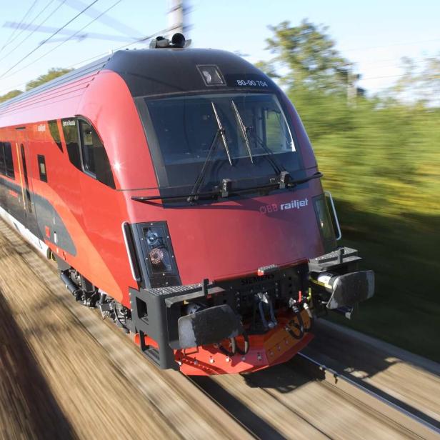 Ein roter ÖBB Railjet-Zug fährt mit hoher Geschwindigkeit durch eine grüne Landschaft.