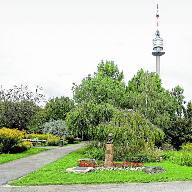Eine Büste steht in einem grünen Park vor dem Donauturm in Wien, umgeben von Bäumen, Blumenbeeten und einem Spazierweg.