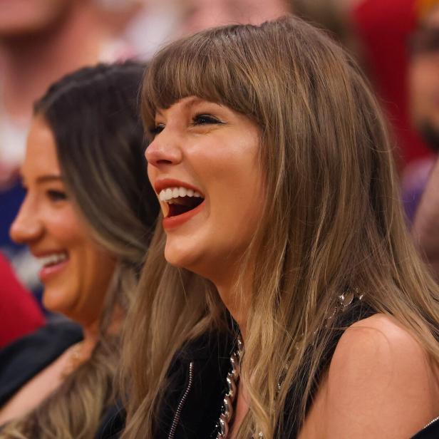 Taylor Swift lacht herzlich, während sie neben einer anderen Frau in einer Menschenmenge sitzt.