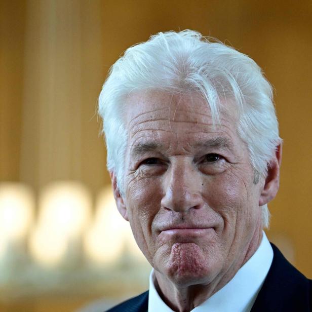 Richard Gere mit weißem Haar und einem sanften Lächeln vor einem unscharfen Hintergrund.