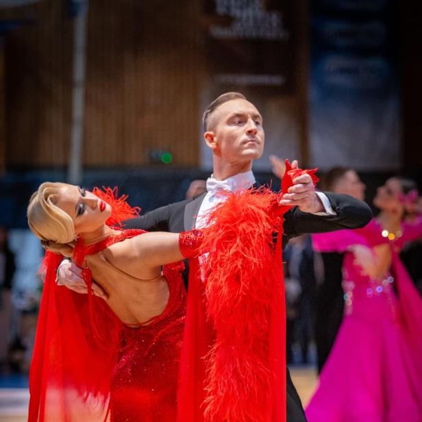 Ein elegantes Tanzpaar tanzt in festlicher Kleidung, die Frau trägt ein rotes Kleid mit Federn.