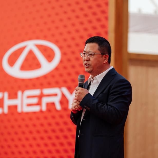 Ein Mann im Anzug spricht mit Mikrofon vor einer roten Wand mit dem Chery-Logo und Schriftzug.
