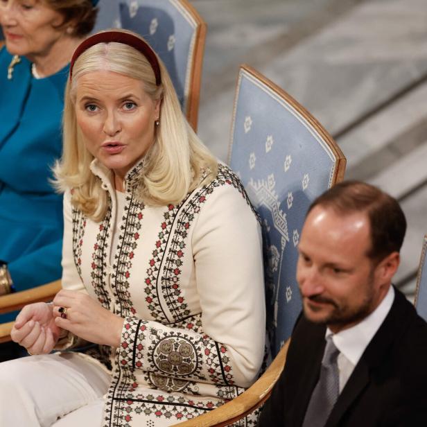 Kronprinzessin Mette-Marit von Norwegen sitzt neben Kronprinz Haakon in einem hellen, bestickten Mantel auf einem blauen Stuhl.