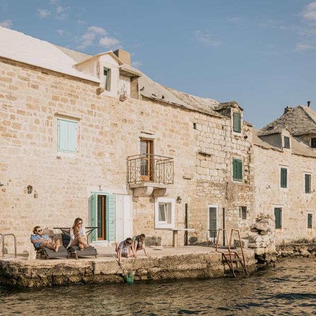 Menschen entspannen sich auf einer Terrasse direkt am Wasser vor einem traditionellen Steinhaus an der kroatischen Adriaküste.