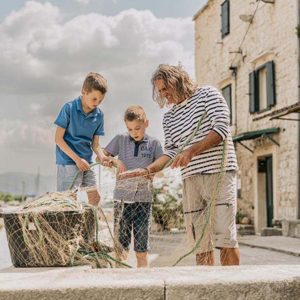 Ein kroatischer Gastgeber zeigt zwei Jungen am Hafen, wie man ein Fischernetz vorbereitet, und vermittelt so lokale Traditionen.