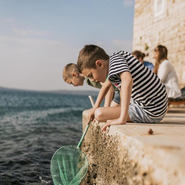 Zwei Kinder fangen am Ufer eines kroatischen Küstendorfs mit einem Kescher im Meer, während Erwachsene im Hintergrund sitzen und die Atmosphäre genießen.