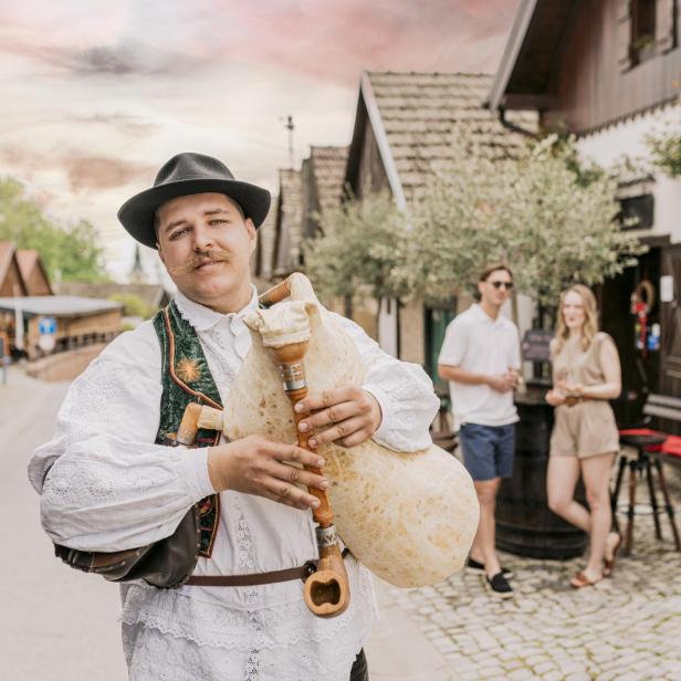 Ein Mann in traditioneller kroatischer Kleidung spielt Dudelsack in einem historischen Dorf, während zwei junge Gäste im Hintergrund stehen.