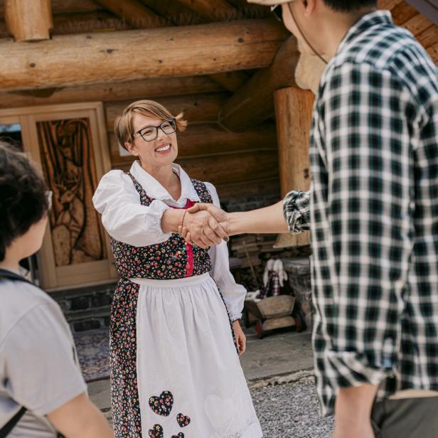 Eine Gastgeberin in traditioneller Tracht begrüßt Gäste herzlich vor einer rustikalen Unterkunft in Gorski Kotar, Kroatien.