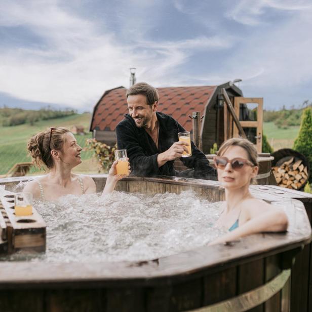 Drei Personen entspannen in einem hölzernen Whirlpool im Freien und stoßen mit Getränken an, während im Hintergrund eine Fasssauna zu sehen ist.