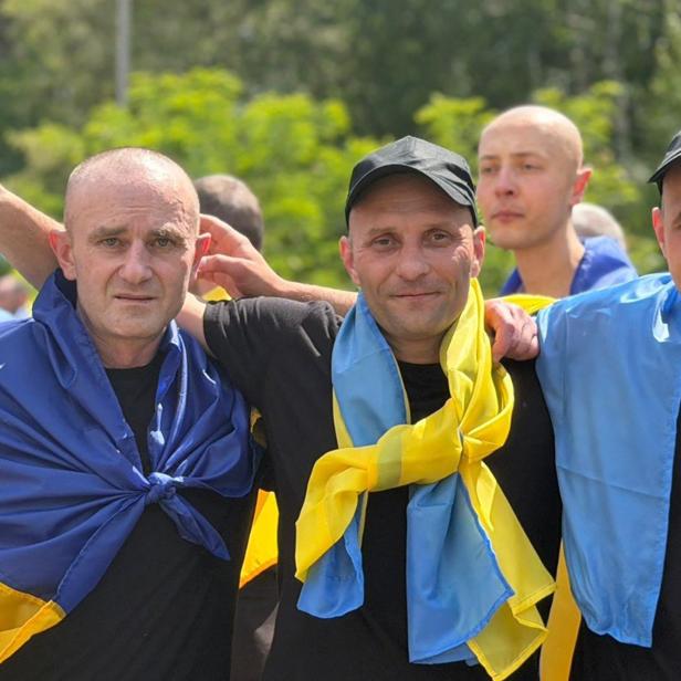 Männer, die in eine ukrainische Fahne gehüllt sind, legen sich die Arme um die Schultern und blicken in die Kamera.