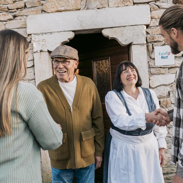 Ein älteres Gastgeberpaar begrüßt zwei Gäste herzlich vor einem Steinhaus mit Local Host-Schild, die Gastgeberin trägt traditionelle kroatische Kleidung.