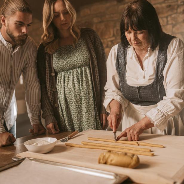 Eine Gastgeberin in traditioneller Kleidung zeigt zwei Gästen, wie man nach Familienrezept Pasta zubereitet.