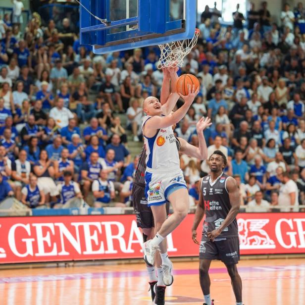 Ein Basketballspieler im weißen Trikot springt zum Korbleger, während ein Verteidiger im schwarzen Trikot versucht zu blocken.