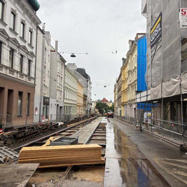 Straße mit Baustelle und offenen Straßenbahnschienen, umgeben von mehrstöckigen Häusern und Baugerüsten bei nassem Wetter.