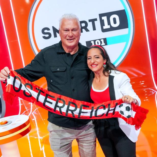 Anton Polster und Caroline Athanasiadis halten gemeinsam einen rot-weiß-roten Schal mit der Aufschrift „Österreich“ vor einer bunten Spielshow-Kulisse.