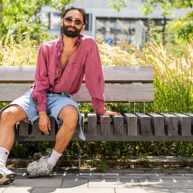 Ein Mann mit Sonnenbrille, offenem roten Hemd und Jeansshorts sitzt entspannt auf einer Holzbank im Grünen.