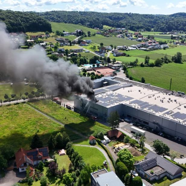 Dichter schwarzer Rauch steigt aus einem brennenden Industriegebäude in ländlicher Umgebung auf, Feuerwehr ist im Einsatz.