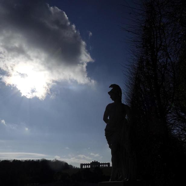 Die Silhouette einer antiken Statue steht vor der Gloriette im Schlosspark Schönbrunn unter einem bewölkten Himmel.