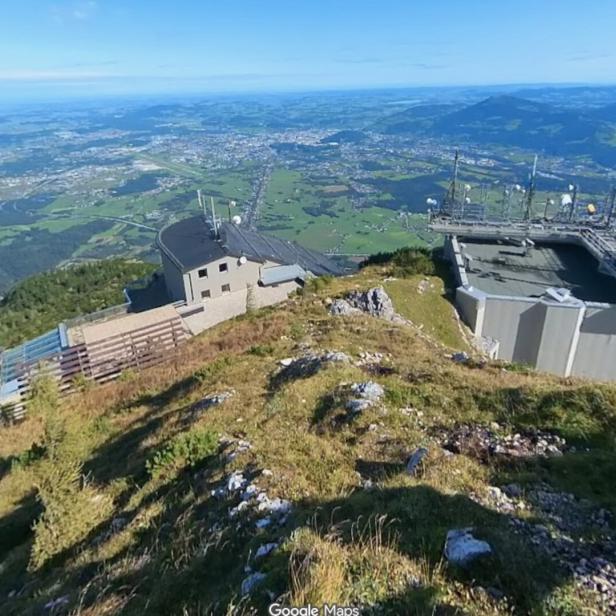 Gebäude mit Antennen auf einem bewachsenen Bergrücken, im Hintergrund weite grüne Landschaft und Berge unter blauem Himmel.
