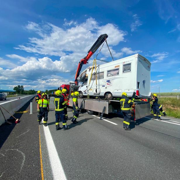 Mehrere Feuerwehrleute bergen mit einem Kran ein beschädigtes Wohnmobil auf einer Autobahn.