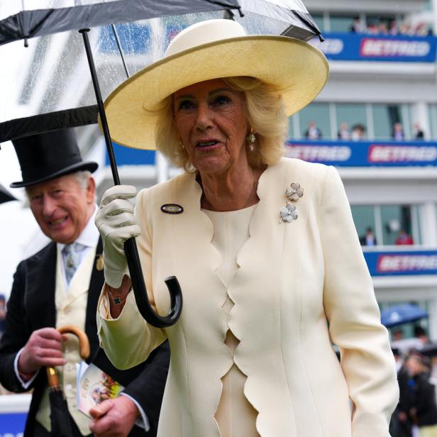 Königin Camilla hält bei Regen einen Regenschirm, während König Charles III. im Hintergrund bei einem Pferderennen steht.