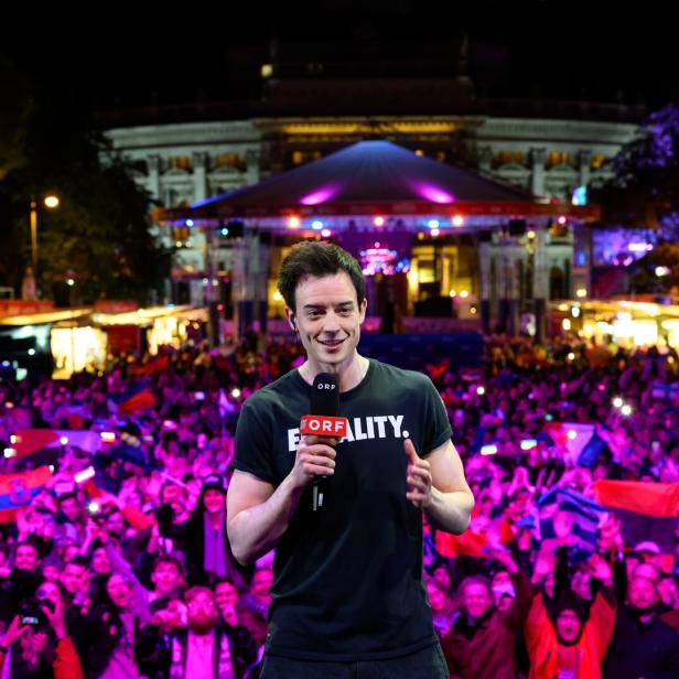 Ein Mann spricht mit einem Mikrofon vor einer jubelnden Menschenmenge bei einer nächtlichen Open-Air-Veranstaltung.