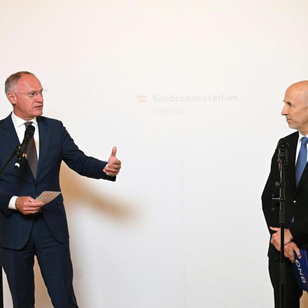Zwei Männer im Anzug stehen vor Mikrofonen und sprechen miteinander vor einer weißen Wand mit Behördenaufschrift.