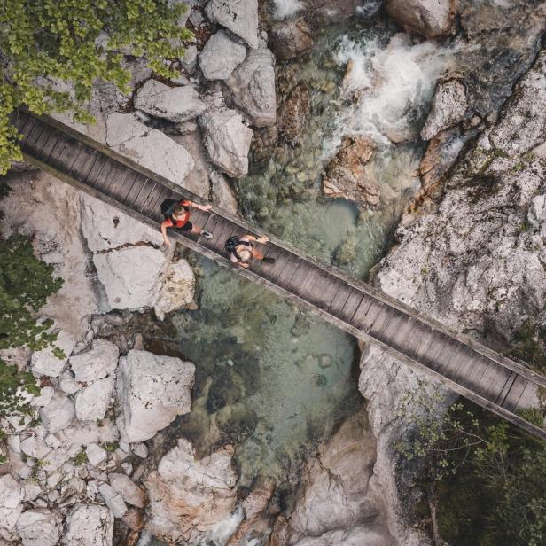 Luftaufnahme einer schmalen Holzbrücke über einen felsigen Bach mit zwei darauf sitzenden Personen und umgebendem Grün.