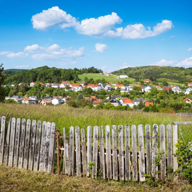 Ein altes Holzzaun steht vor einer Wiese, dahinter ein Dorf mit roten Dächern und grünen Hügeln unter blauem Himmel.
